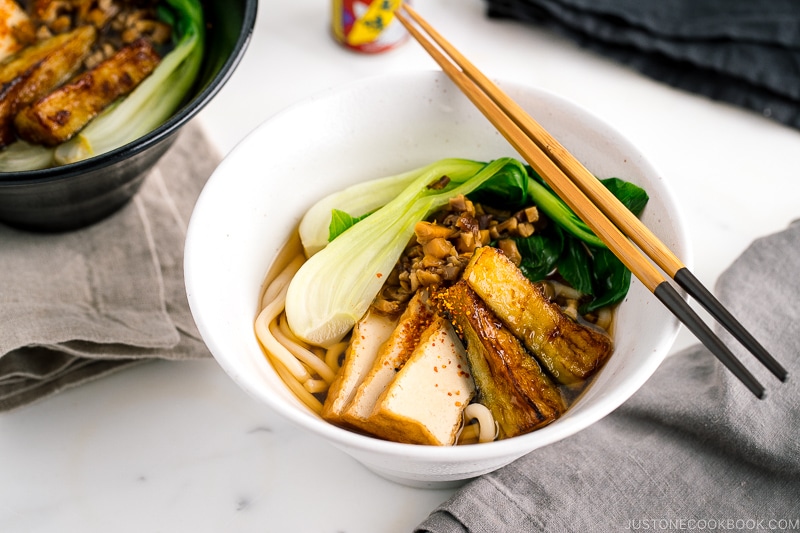 A white bowl containing Vegetarian Udon Noodle Soup topped with bok choy, fried tofu, minced mushroom, and crispy eggplant.