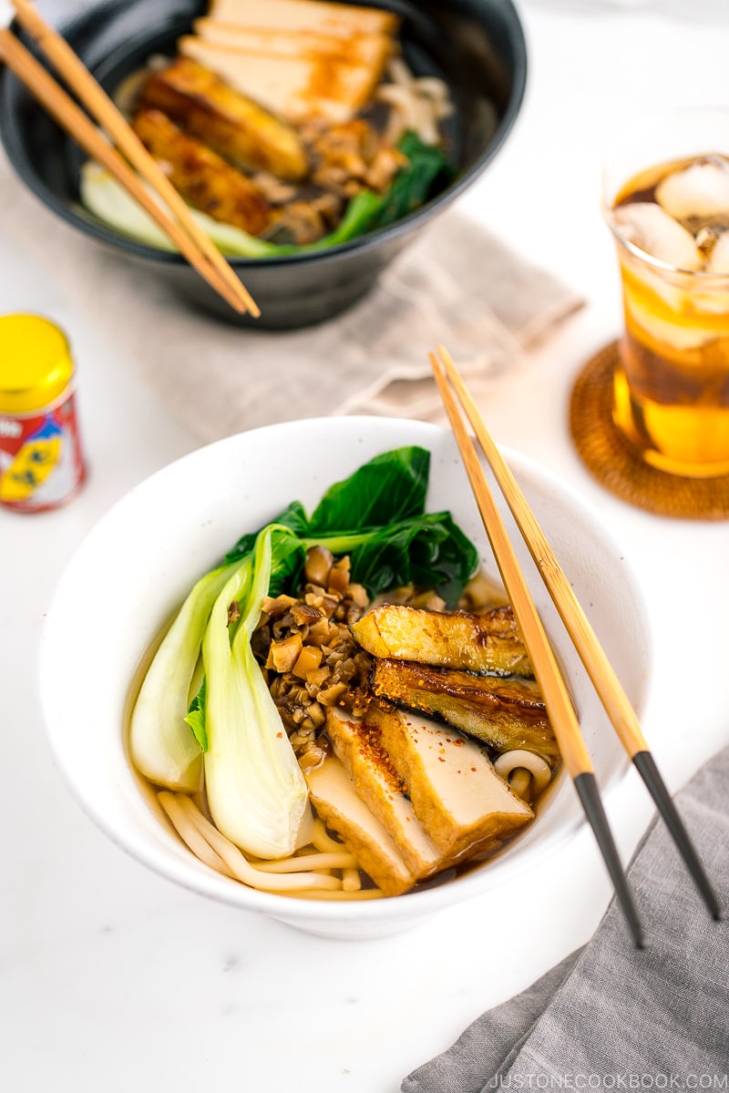 Un bol blanc contenant une soupe végétarienne aux nouilles Udon garnie de bok choy, de tofu frit, de champignons émincés et d'aubergines croustillantes.