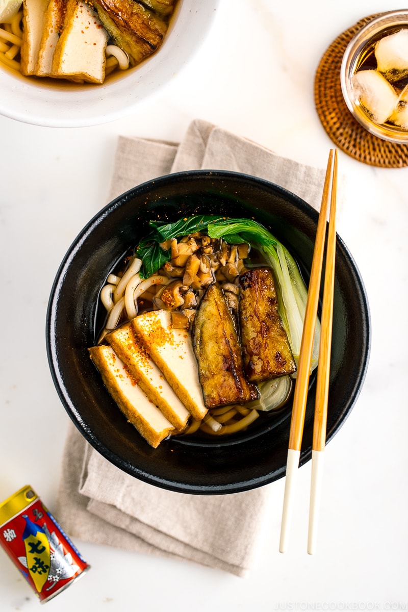 A black bowl containing Vegetarian Udon Noodle Soup topped with bok choy, fried tofu, minced mushroom, and crispy eggplant.