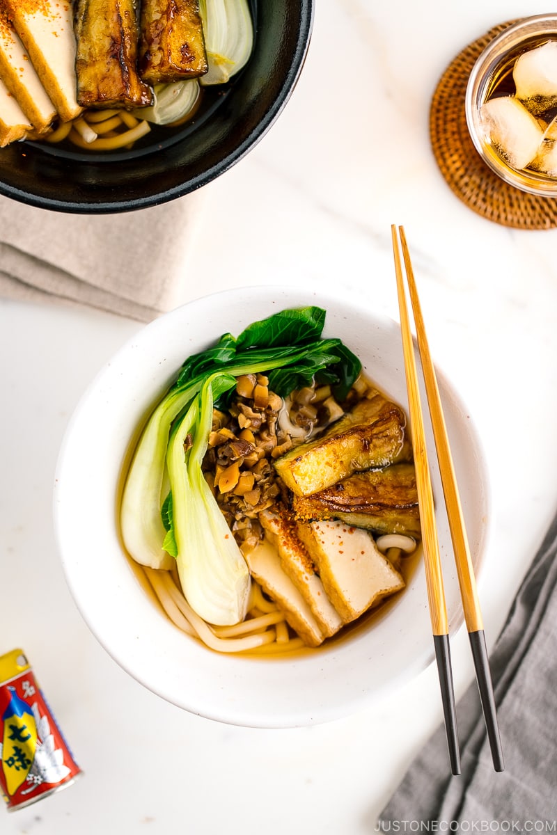 A white bowl containing Vegetarian Udon Noodle Soup topped with bok choy, fried tofu, minced mushroom, and crispy eggplant.