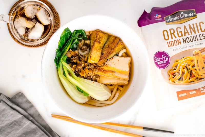 A white bowl containing Vegetarian Udon Noodle Soup topped with bok choy, fried tofu, minced mushroom, and crispy eggplant.