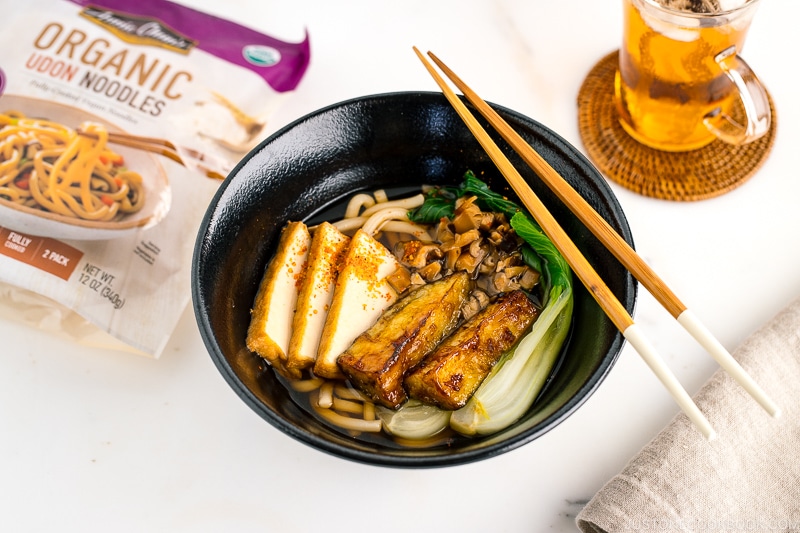 A black bowl containing Vegetarian Udon Noodle Soup topped with bok choy, fried tofu, minced mushroom, and crispy eggplant.