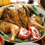 A white platter containing Honey Soy Glazed Spatchcock Chicken decorated with sage, pomegranate, and cranberries.