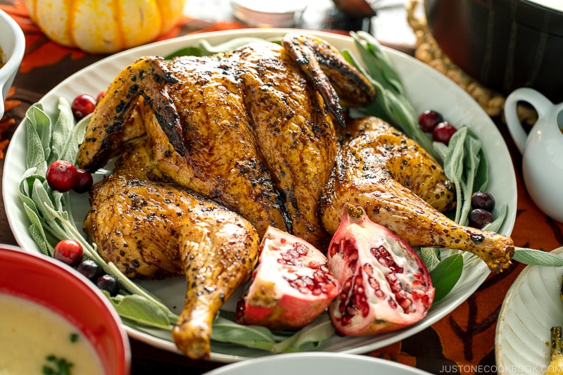 A white platter containing Honey Soy Glazed Spatchcock Chicken decorated with sage, pomegranate, and cranberries.