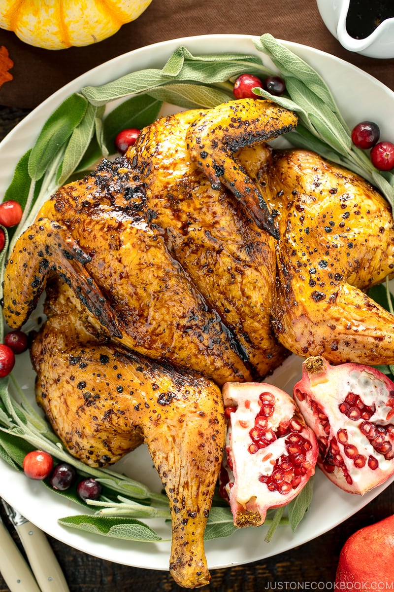 A white platter containing Honey Soy Glazed Spatchcock Chicken decorated with sage, pomegranate, and cranberries.