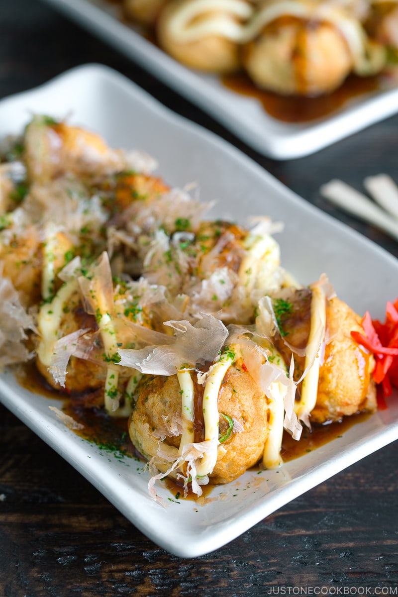 A white plate containing Takoyaki (Octopus Balls) topped with a generous drizzle of takoyaki sauce, Japanese mayo, aonori, katsuobushi.