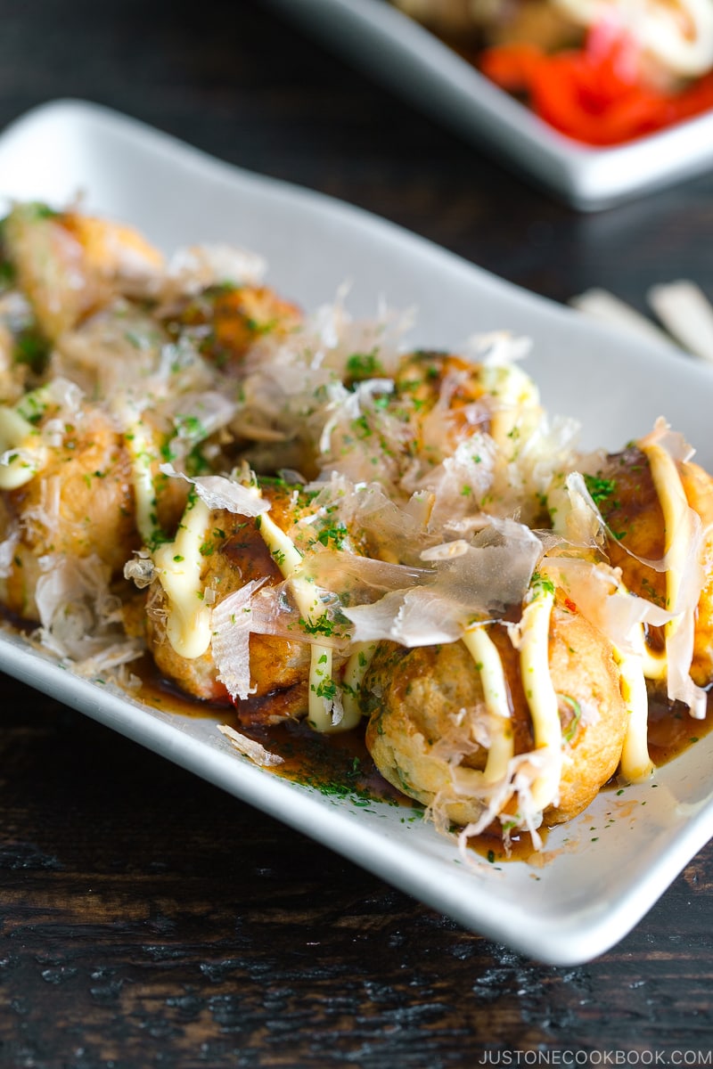 A white plate containing Takoyaki (Octopus Balls) topped with a generous drizzle of takoyaki sauce, Japanese mayo, aonori, katsuobushi.