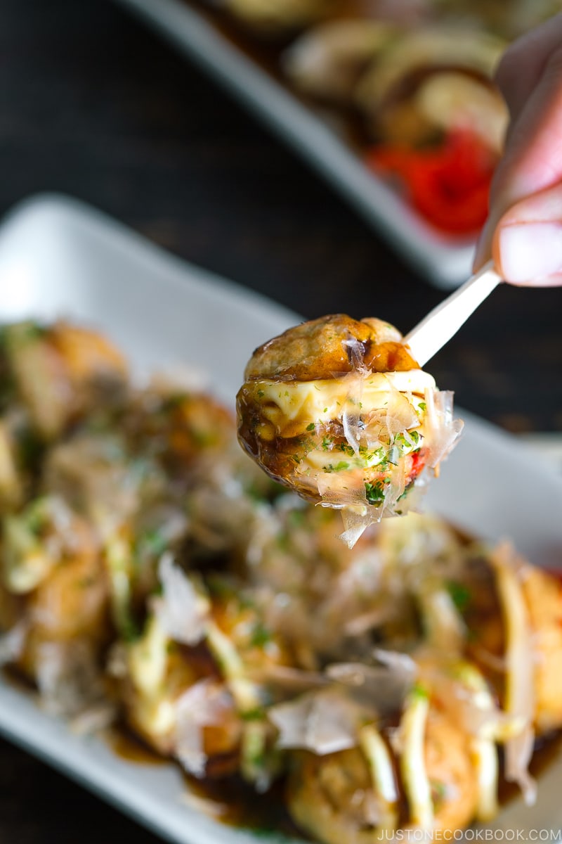 A white plate containing Takoyaki (Octopus Balls) topped with a generous drizzle of takoyaki sauce, Japanese mayo, aonori, katsuobushi.