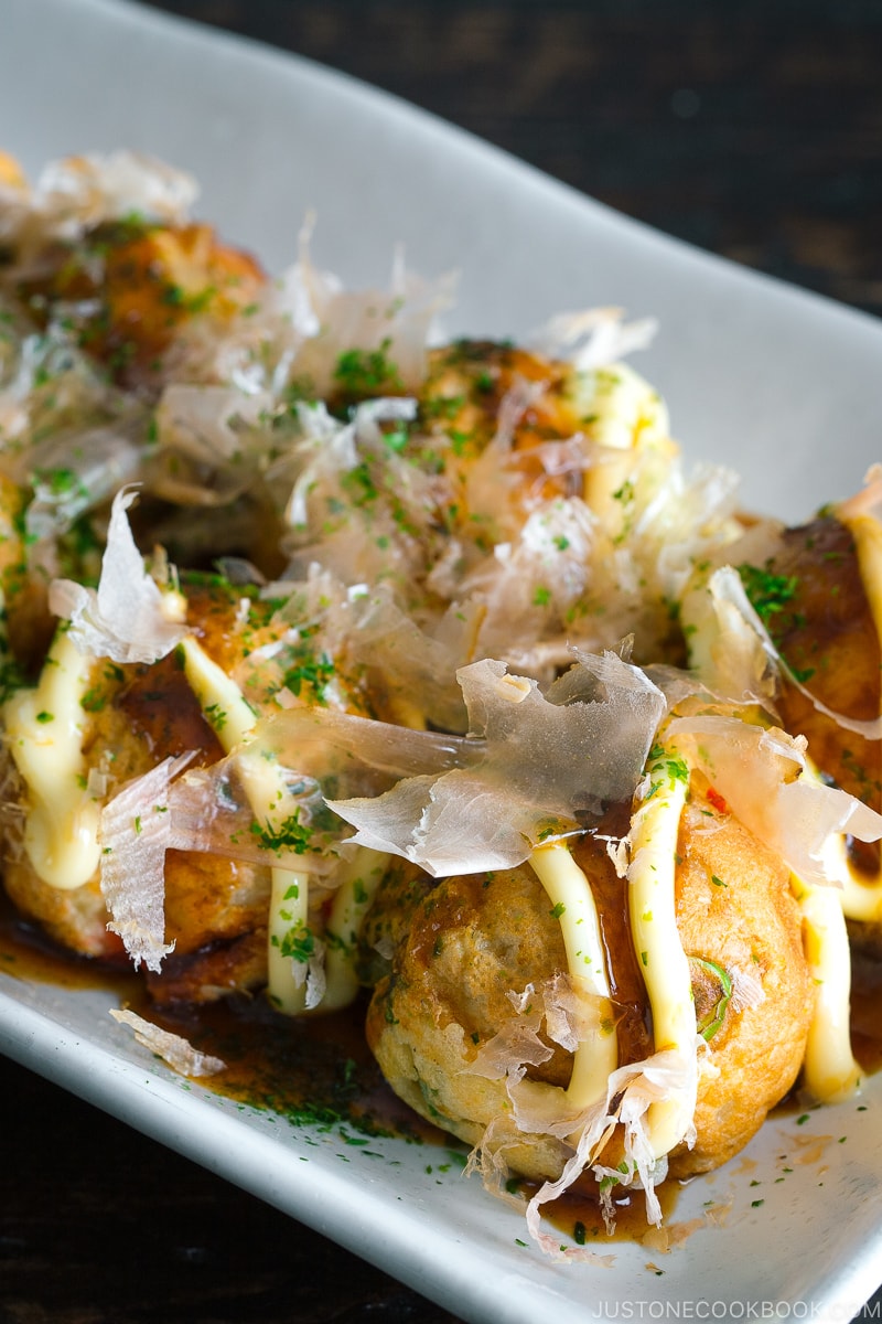 A white plate containing Takoyaki (Octopus Balls) topped with a generous drizzle of takoyaki sauce, Japanese mayo, aonori, katsuobushi.