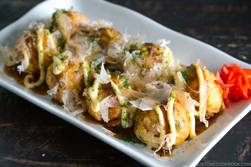 A white plate containing Takoyaki (Octopus Balls) topped with a generous drizzle of takoyaki sauce, Japanese mayo, aonori, katsuobushi.