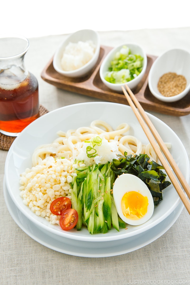 A white bowl containing chilled udon noodles, tenkasu, julienned cucumber, boiled egg, wakame seaweed, and grated onion, along with savory noodle soup.