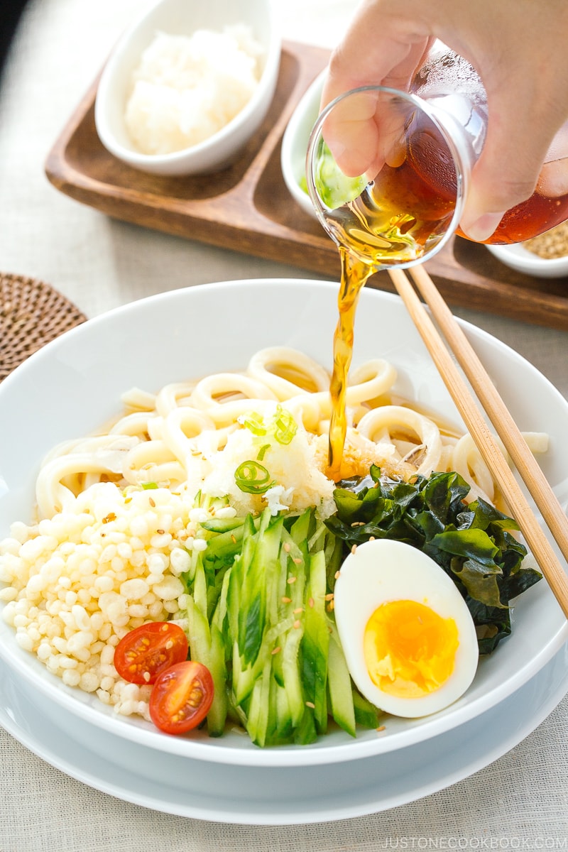 A white bowl containing chilled udon noodles, tenkasu, julienned cucumber, boiled egg, wakame seaweed, and grated onion, along with savory noodle soup.