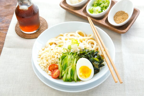A white bowl containing chilled udon noodles, tenkasu, julienned cucumber, boiled egg, wakame seaweed, and grated onion, along with savory noodle soup.