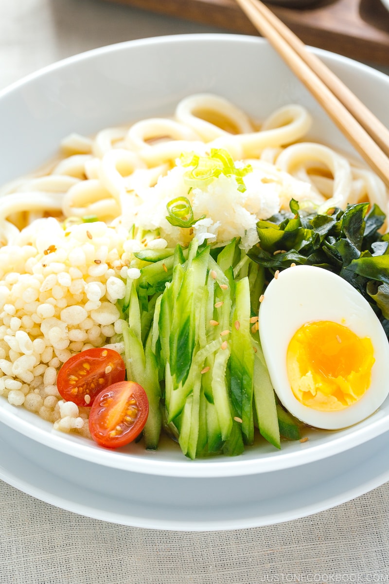 A white bowl containing chilled udon noodles, tenkasu, julienned cucumber, boiled egg, wakame seaweed, and grated onion, along with savory noodle soup.