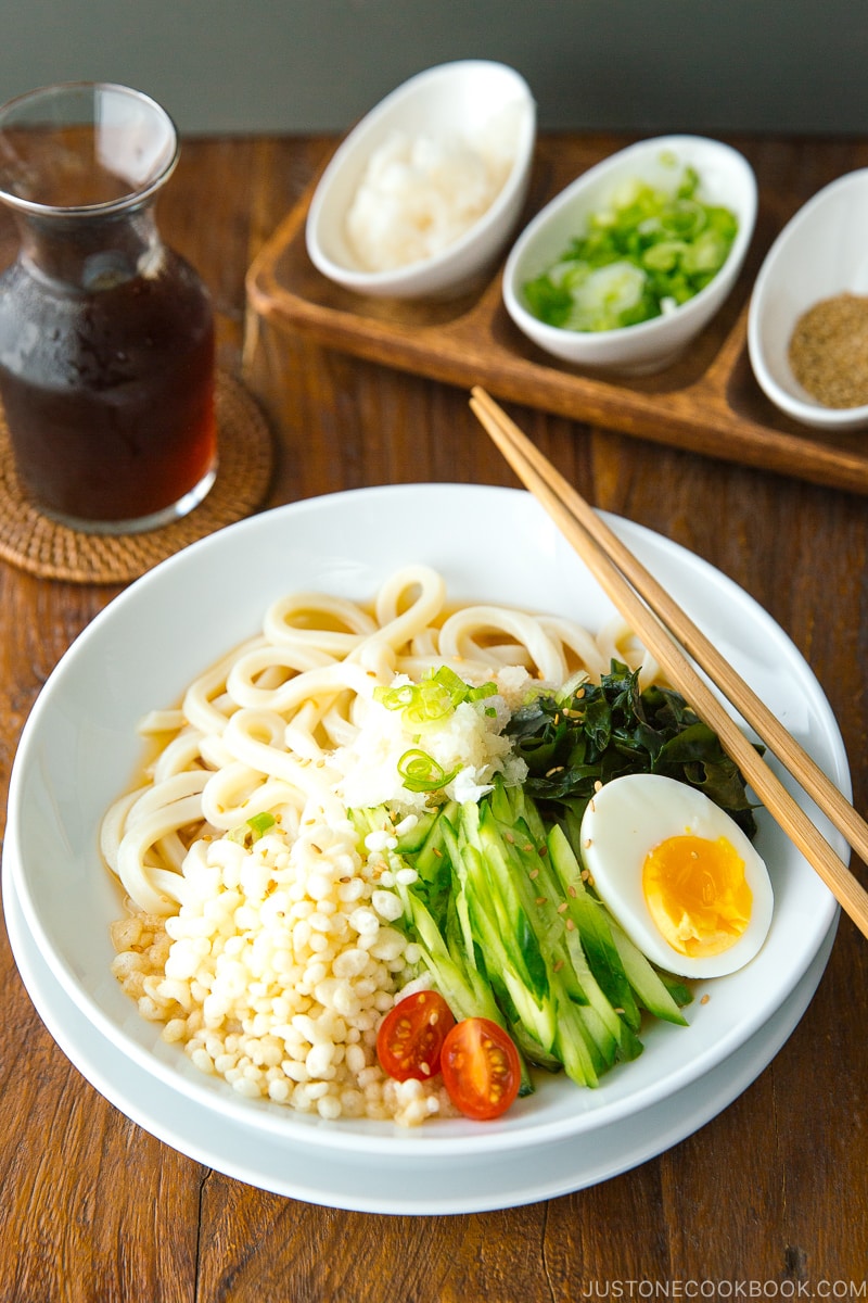 A white bowl containing chilled udon noodles, tenkasu, julienned cucumber, boiled egg, wakame seaweed, and grated onion, along with savory noodle soup.