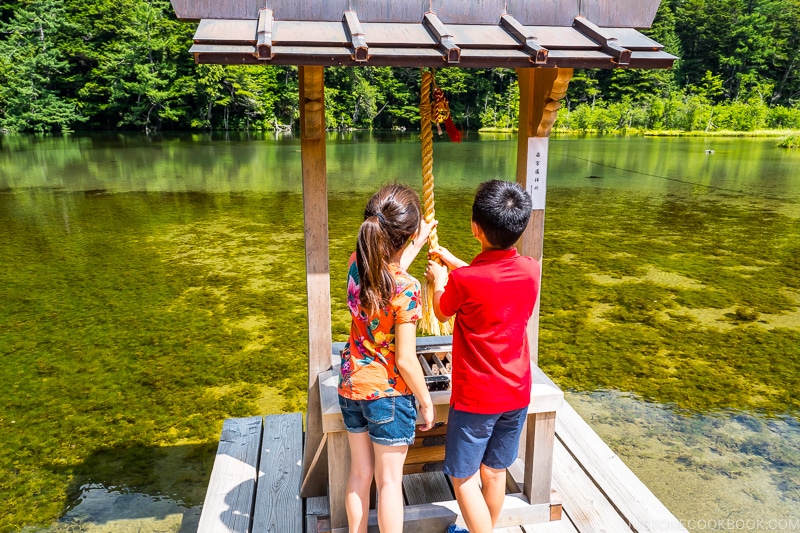 two children pulling on a rope to ring the bell at an altar next to a large pond
