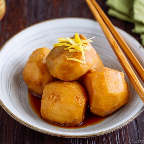 A white ceramic bowl containing simmered taro topped with julienned lemon zest.