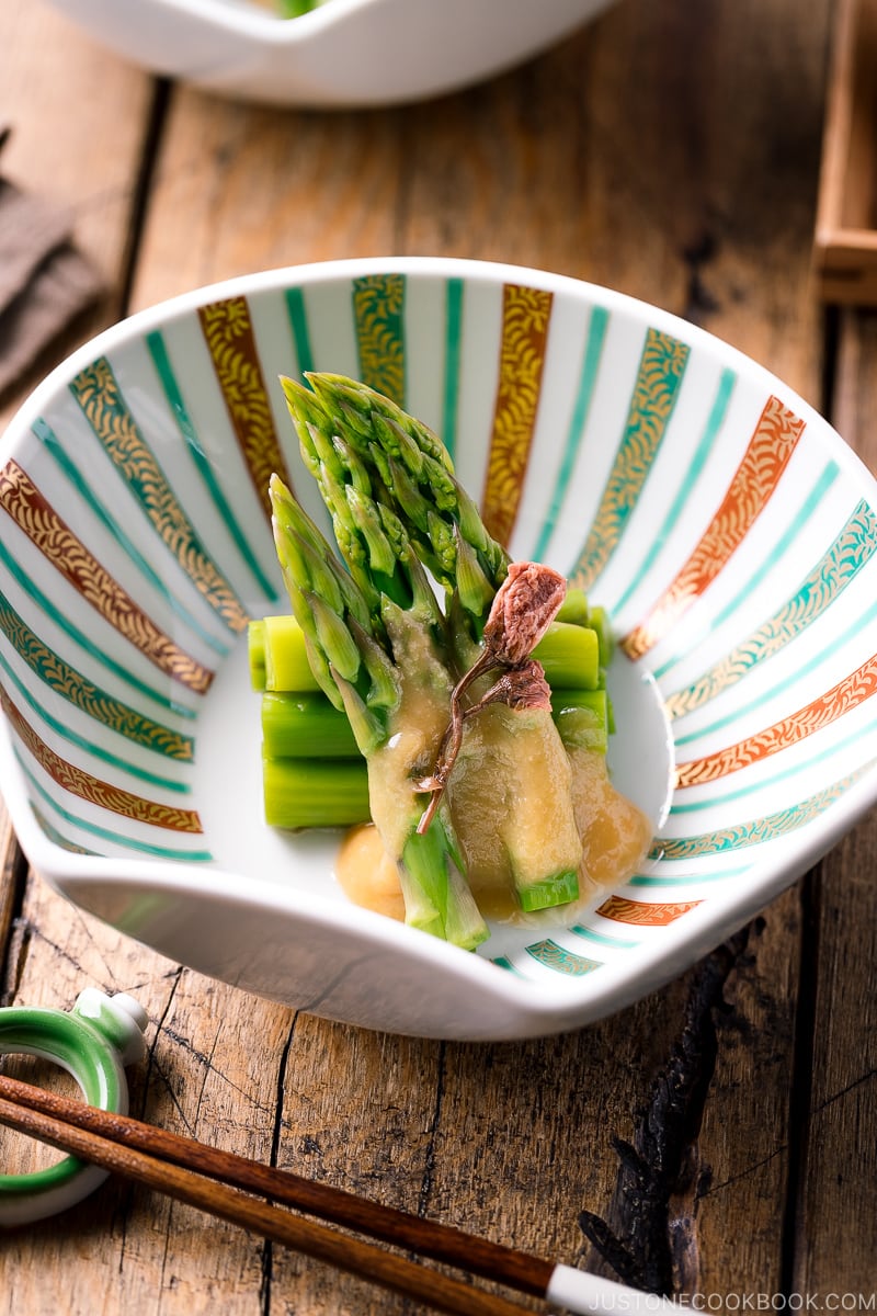 A shell shaped bowl containing Asparagus with Miso Dressing (Karashi Sumisoae) garnished with salt pickled cherry blossom.