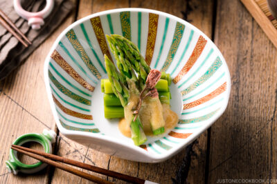 A shell shaped bowl containing Asparagus with Miso Dressing (Karashi Sumisoae) garnished with salt pickled cherry blossom.