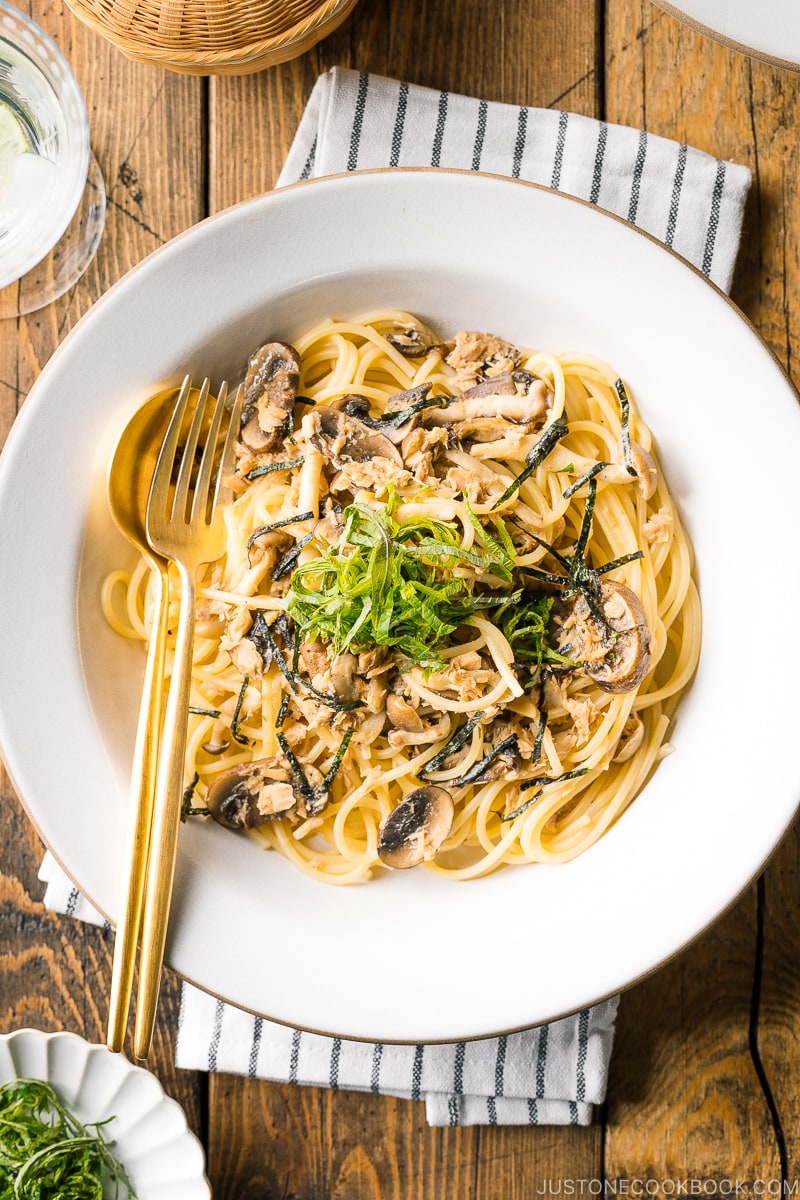 Une assiette en céramique blanche contenant des pâtes au thon et aux champignons à la japonaise garnies de feuilles de nori et de shiso râpées.