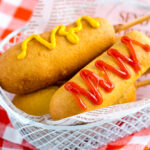 A basket containing corn dogs served with ketchup and mustard drizzle.