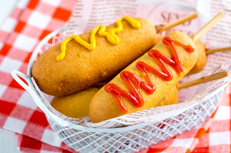 A basket containing corn dogs served with ketchup and mustard drizzle.