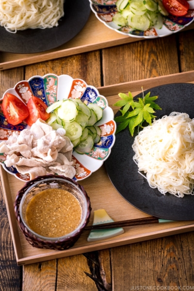 A wooden tray containing a plate of somen noodles, a bowl of sesame miso dipping sauce, and a plate of pork shabu shabu and cucumber slices.