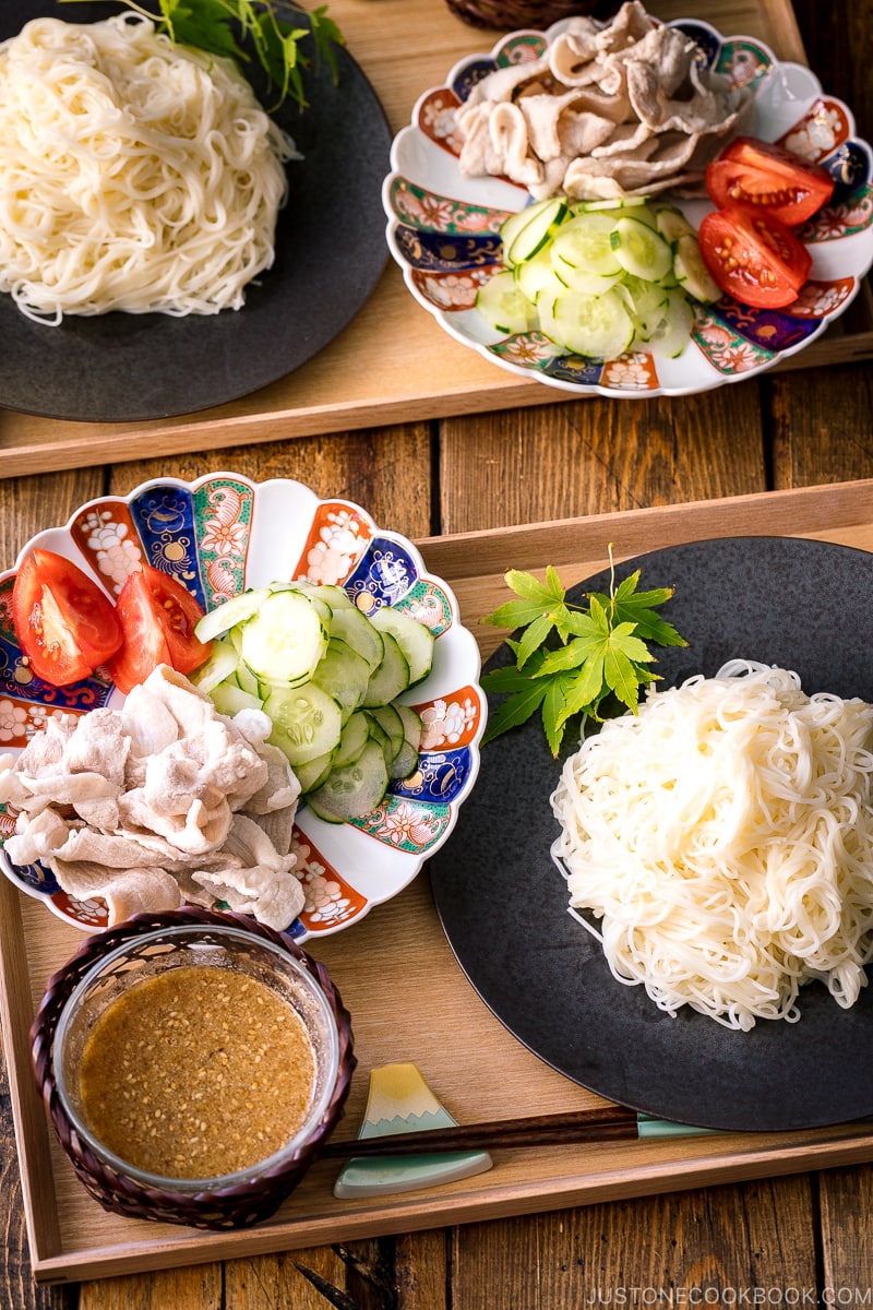 A wooden tray containing a plate of somen noodles, a bowl of sesame miso dipping sauce, and a plate of pork shabu shabu and cucumber slices.