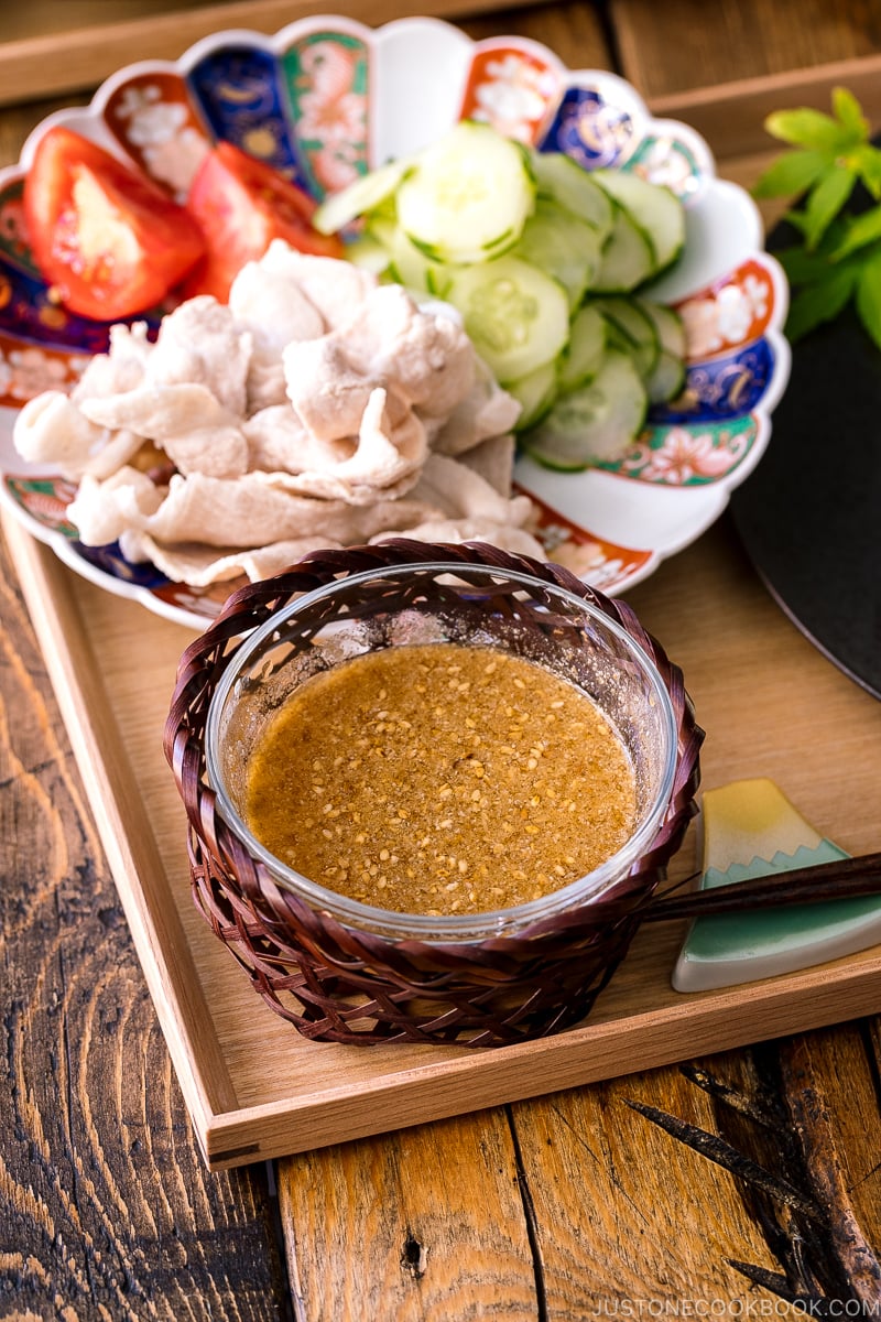 A wooden tray containing a bowl of sesame miso dipping sauce, and a plate of pork shabu shabu and cucumber slices.