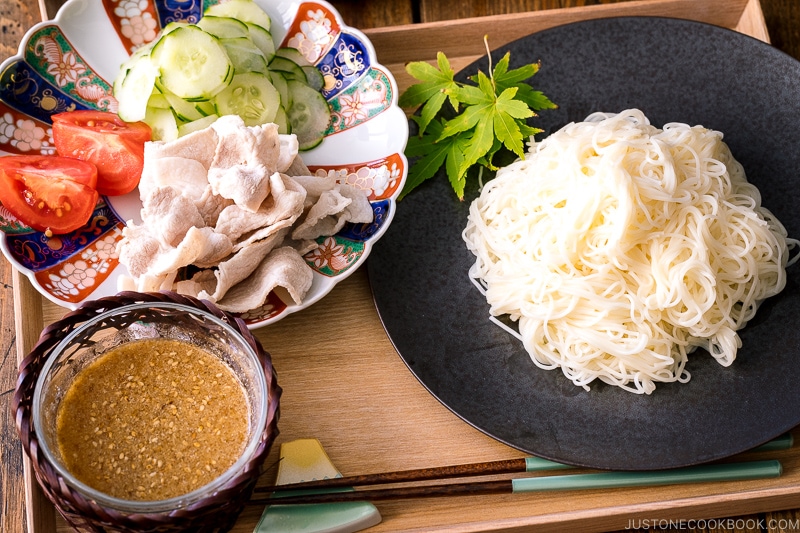 A wooden tray containing a plate of somen noodles, a bowl of sesame miso dipping sauce, and a plate of pork shabu shabu and cucumber slices.