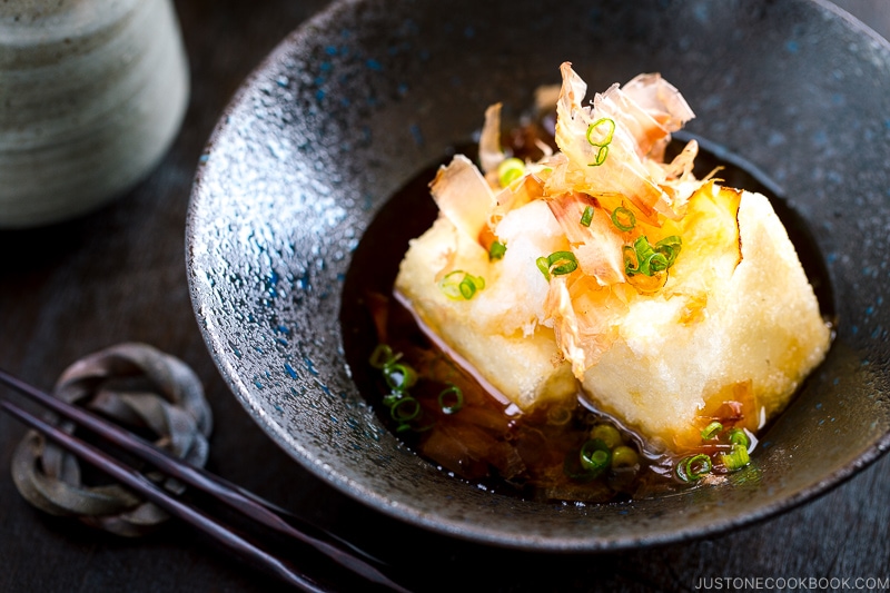 A black ceramic bowl containing Agedashi Tofu sprinkled with katsuobushi and scallion.