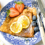 A blue plate containing lemon and sugar crepe along with fresh fruits.