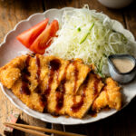 A white plate containing chicken katsu with tonkatsu sauce and shredded cabbage with sesame dressing.