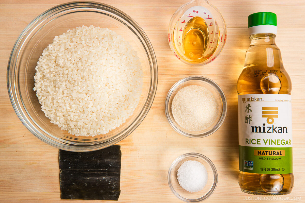 A top-down view of sushi rice ingredients for inari sushi: a bowl of uncooked rice, a measuring cup of rice vinegar, a bottle of rice vinegar, sugar, salt, and dried kombu arranged neatly on a wooden surface.