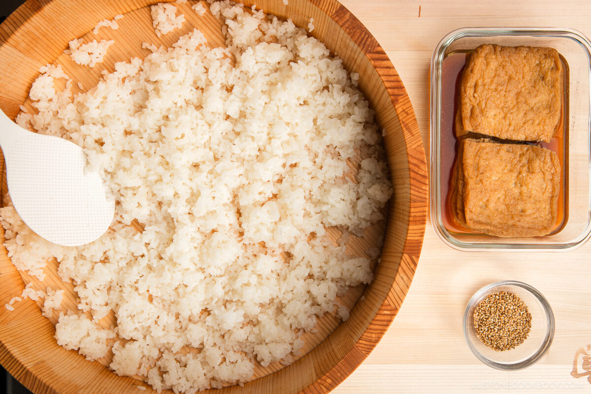 A wooden bowl filled with cooked sushi rice and a white rice paddle sits beside a glass dish of inari sushi&mdash;two pieces of fried tofu in sauce&mdash;and a small bowl of sesame seeds on a light wooden surface.