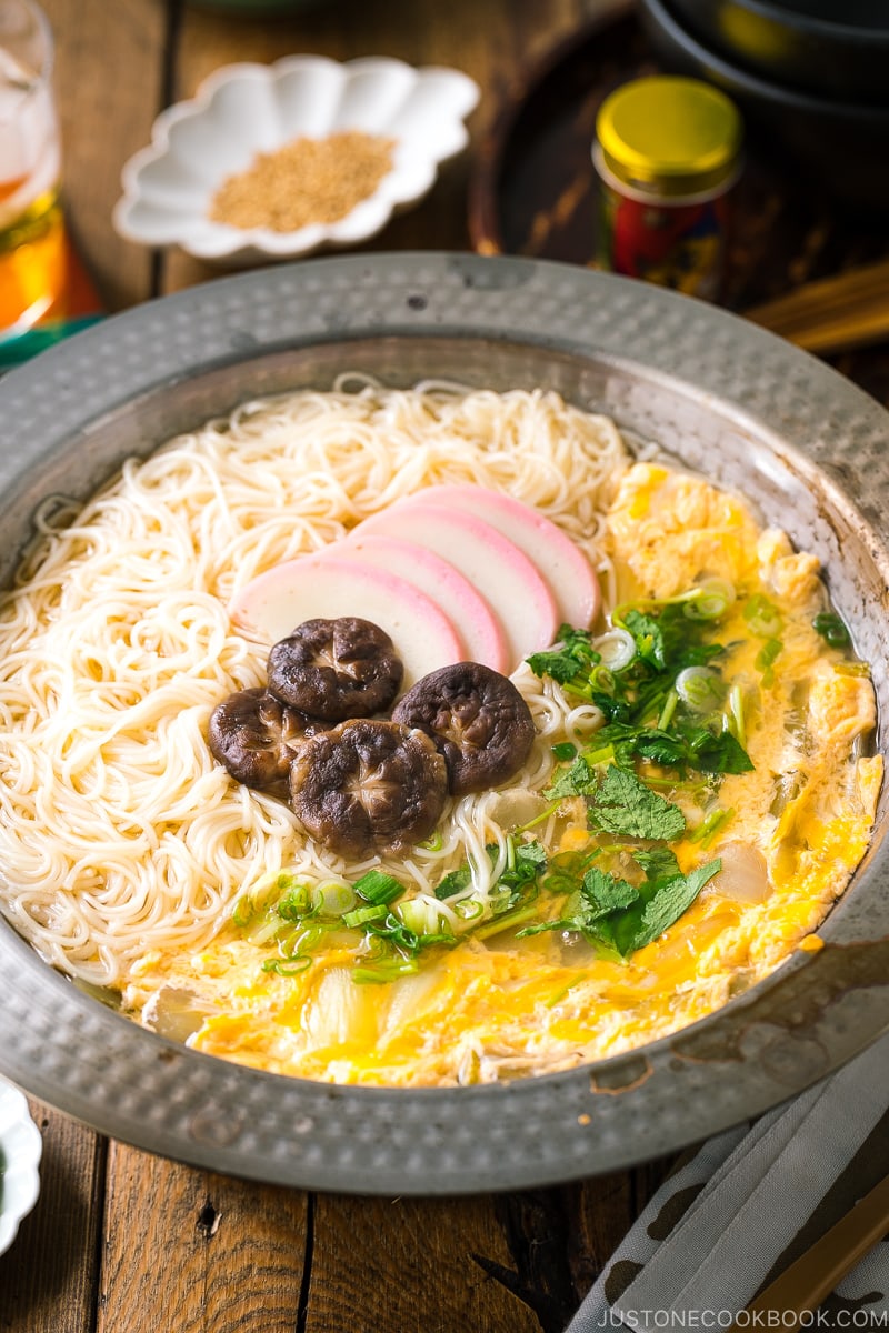 A yukihira nabe pot containing somen noodle soup with vegetables, shiitake mushrooms, egg, kamaboko fish cake, garnish with green onion.