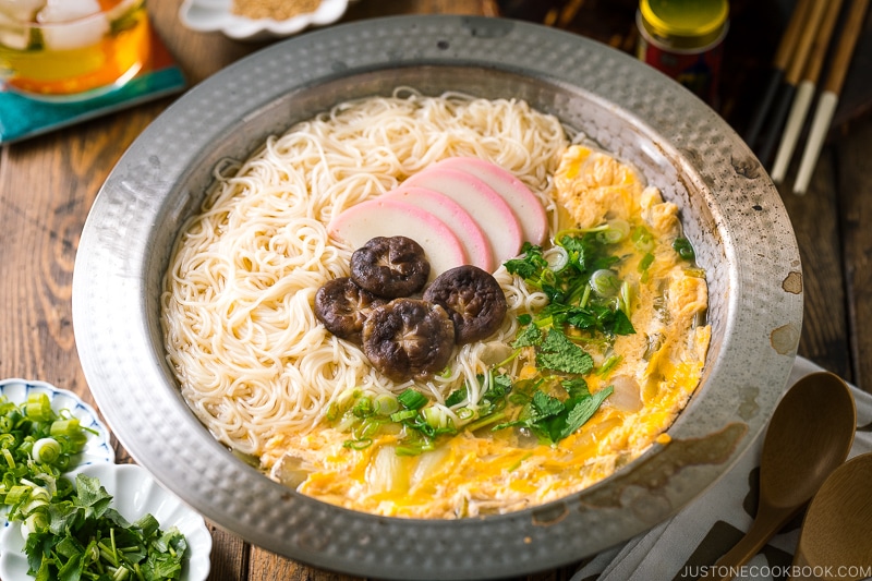 A yukihira nabe pot containing somen noodle soup with vegetables, shiitake mushrooms, egg, kamaboko fish cake, garnish with green onion.