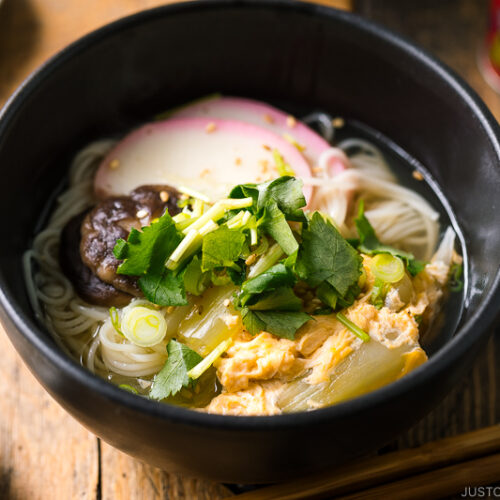 A black bowl containing somen noodle soup with vegetables, shiitake mushrooms, egg, kamaboko fish cake, garnish with green onion.