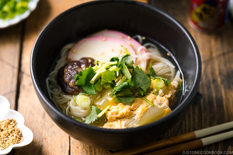 A black bowl containing somen noodle soup with vegetables, shiitake mushrooms, egg, kamaboko fish cake, garnish with green onion.