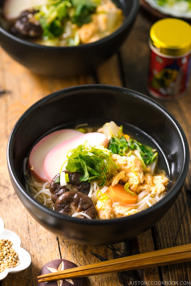 A black bowl containing somen noodle soup with vegetables, shiitake mushrooms, egg, kamaboko fish cake, garnish with green onion.