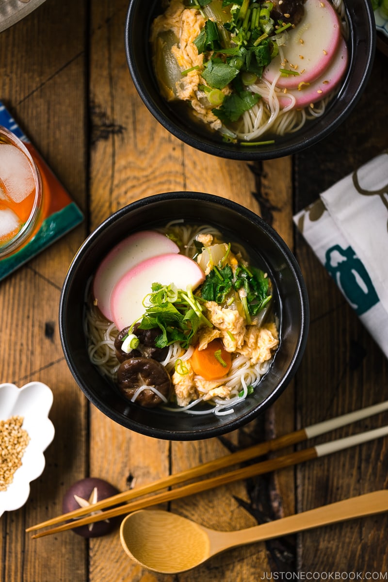 A black bowl containing somen noodle soup with vegetables, shiitake mushrooms, egg, kamaboko fish cake, garnish with green onion.