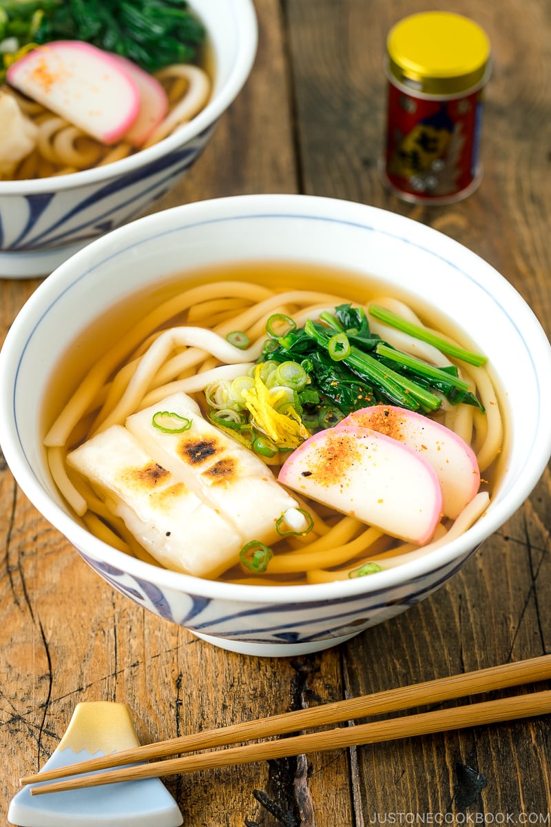 A donburi bowl containing udon noodles topped with toasted mochi, spinach, and fish cake.