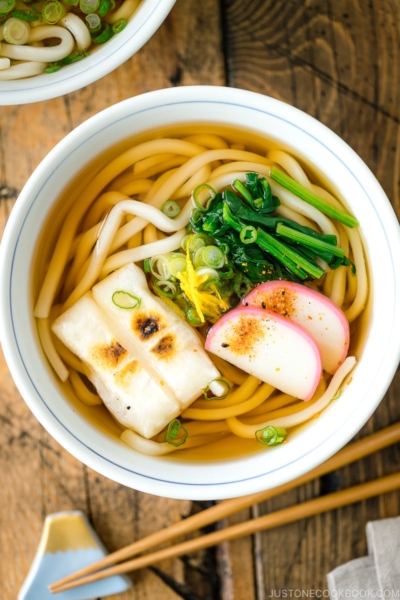 A donburi bowl containing udon noodles topped with toasted mochi, spinach, and fish cake.