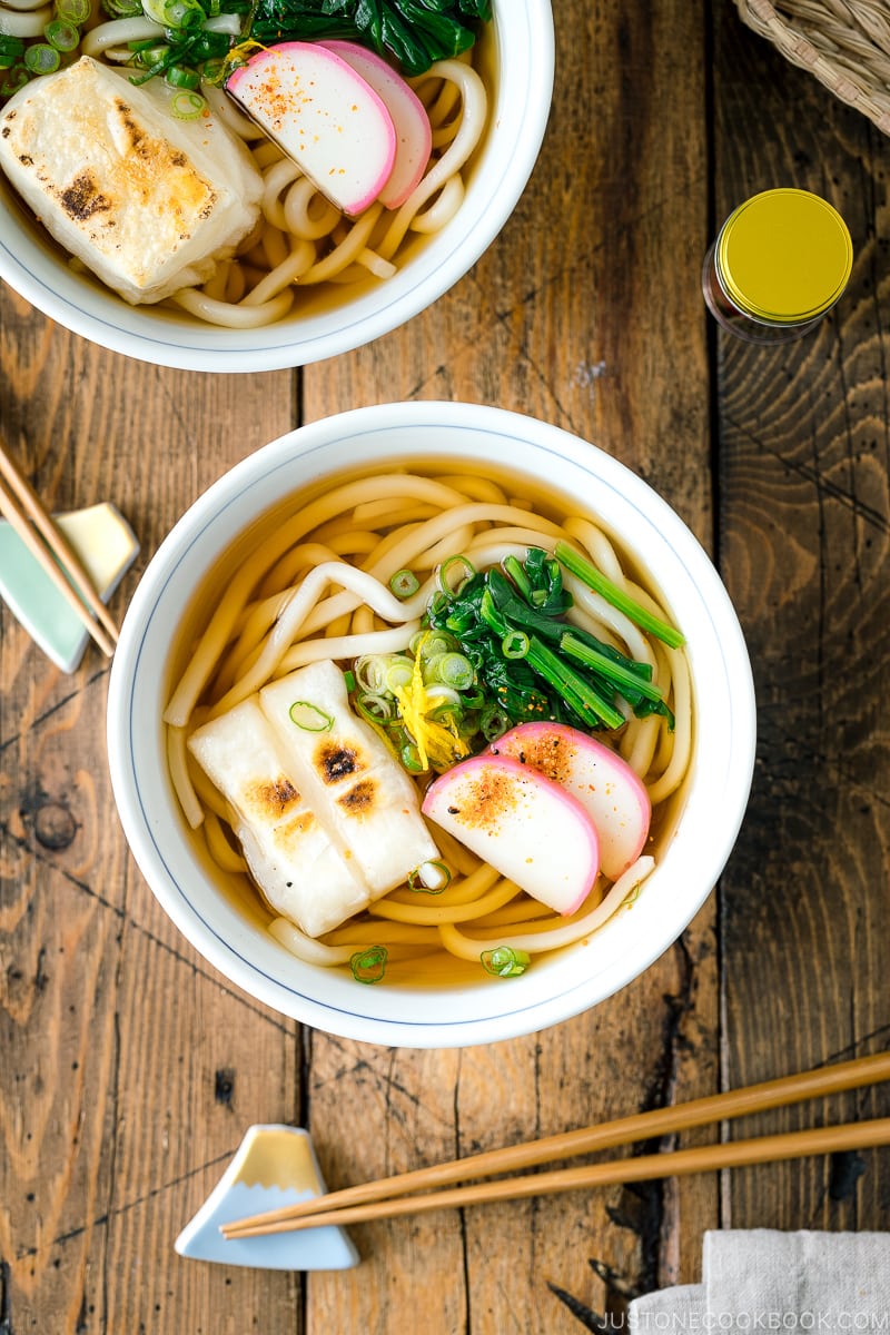 A donburi bowl containing udon noodles topped with toasted mochi, spinach, and fish cake.