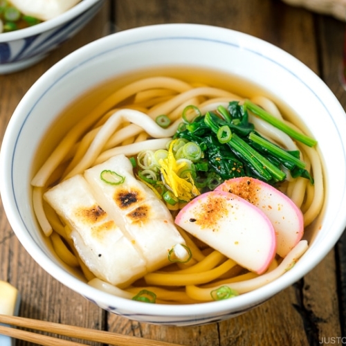 A donburi bowl containing udon noodles topped with toasted mochi, spinach, and fish cake.