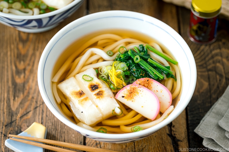 A donburi bowl containing udon noodles topped with toasted mochi, spinach, and fish cake.