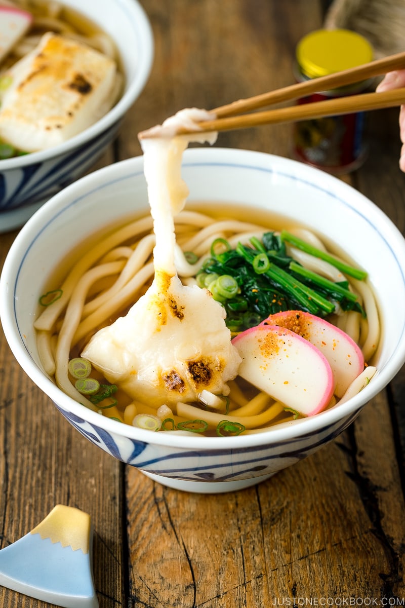 A donburi bowl containing udon noodles topped with toasted mochi, spinach, and fish cake.