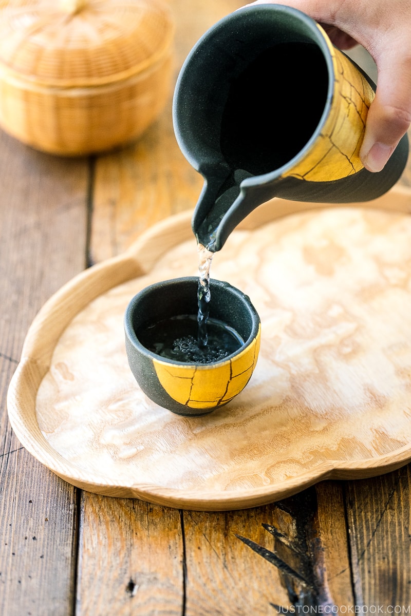 sake being served from a black vessel into a cup on a wooden tray