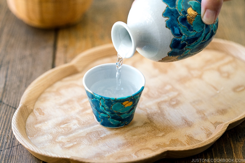 sake being served from a white vessel into a cup on a wooden tray