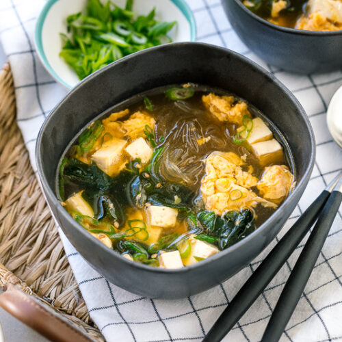 Black ceramic bowls containing glass noodle soup with tofu, fluffy egg, and wakame seaweed.